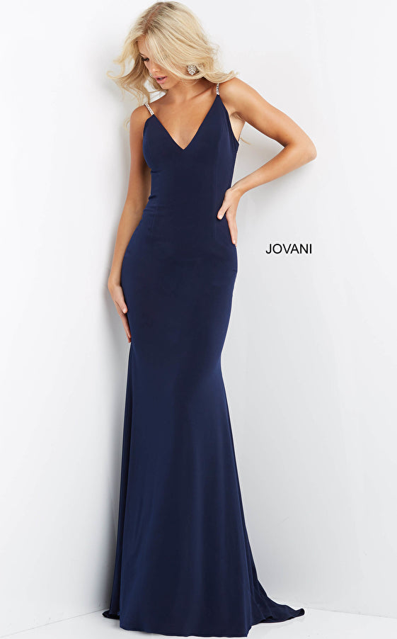 A woman in a fitted, navy blue Jovani Prom Gown 07297 with thin straps and a deep V-neckline stands against a white background. The elegant backless mermaid dress features
